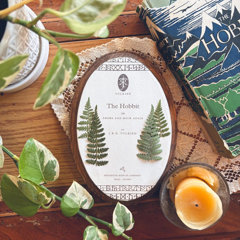 May include: An oval wooden frame with the book cover of "The Hobbit" by J.R.R. Tolkien. The book cover is white with green ferns and text. A vintage copy of the book is visible in the background, along with a lit candle and greenery.
