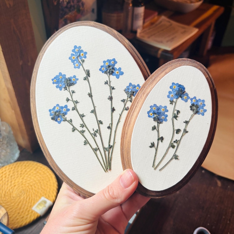 May include: Two oval-shaped wooden frames containing pressed blue forget-me-not flowers on a white fabric background. The larger frame on the left displays a taller arrangement of stems, while the smaller frame on the right features a shorter cluster.