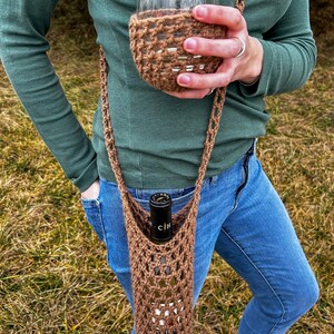 Op de afbeelding: Een bruine gehaakte wijnflesdrager met een band en een bijpassende gehaakte wijnbekerhouder. De drager wordt over de schouder gedragen en houdt een fles wijn vast.