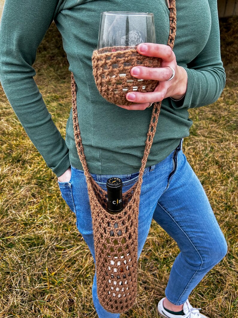 Op de afbeelding: Een bruine gehaakte wijnflesdrager met een band en een bijpassende gehaakte wijnbekerhouder. De drager wordt over de schouder gedragen en houdt een fles wijn vast.