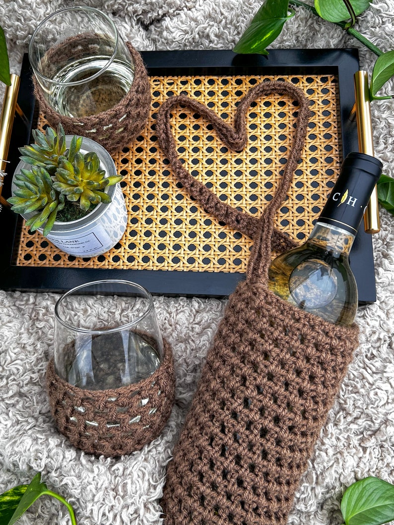 Op de afbeelding: Een bruine gehaakte wijnfleshouder met een hartvormige opening. De houder staat op een gevlochten dienblad met een glas water en een potplant. Een fles witte wijn zit in de houder.