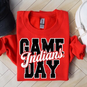 Könnte beinhalten: Ein rotes Sweatshirt mit den Worten "GAME DAY Indians" in einem schwarz-weißen Design. Eine schwarze Baseballkappe und ein weißer Rucksack mit Sonnenbrille sind ebenfalls im Bild.