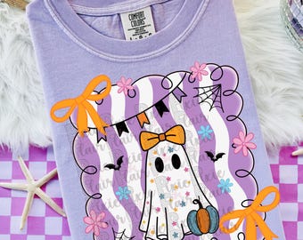 May include: A light purple t-shirt with a Comfort Colors label at the neck. The graphic features a white ghost with an orange bow, holding a blue and orange pumpkin. The design includes orange ribbons, pink flowers, black bats, and spider webs.