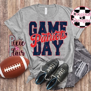 Puede incluir: Una camiseta gris con las palabras "GAME DAY Patriots" en letras azul marino y rojo. La imagen incluye un balón de fútbol americano marrón, zapatillas negras y vaqueros desgastados, todo sobre una superficie de madera.