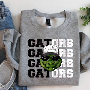 May include: A grey sweatshirt with a graphic of a green alligator wearing sunglasses and a hat. The word "Gators" is printed in a stacked design in black and white. The sweatshirt is paired with blue jeans and grey shoes.