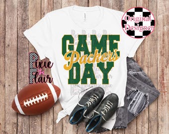 May include: White t-shirt with the text "GAME DAY Packers" in green and yellow. A brown football, black shoes, and distressed jeans are arranged on a wooden surface, suggesting a sports theme.