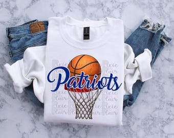 May include: White crew neck sweatshirt featuring a basketball graphic with the word "Patriots" in blue script. The basketball is orange and brown, positioned above a net. The sweatshirt is laid on top of blue jeans.