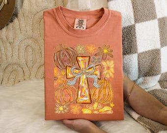 May include: A folded orange t-shirt featuring a cross with a bow, pumpkins, and flowers. The cross is the focal point, with a silver bow. The pumpkins and flowers are in shades of orange and yellow. The shirt is held by a person.