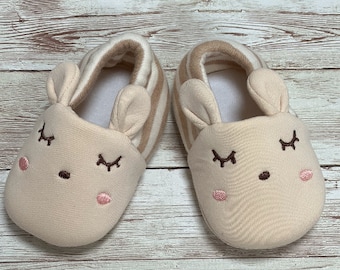 May include: A pair of cream-colored baby booties with brown and white striped cuffs. The booties have embroidered eyes, a nose, and pink cheeks, resembling a cute animal face.