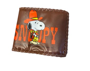 May include: Brown leather wallet featuring Snoopy in a cowboy hat with the word "SNOOPY" in orange. The wallet has a stitched border and a copyright notice at the bottom. A classic Peanuts collectible.