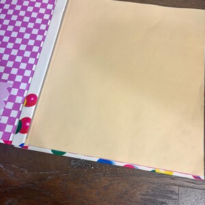 Puede incluir: Un &aacute;lbum de recortes abierto con un interior de color beige claro y una cubierta decorativa. La cubierta presenta un patr&oacute;n de cuadros morados y blancos, con lunares coloridos y un lomo blanco. El &aacute;lbum de recortes est&aacute; sobre una superficie de madera.