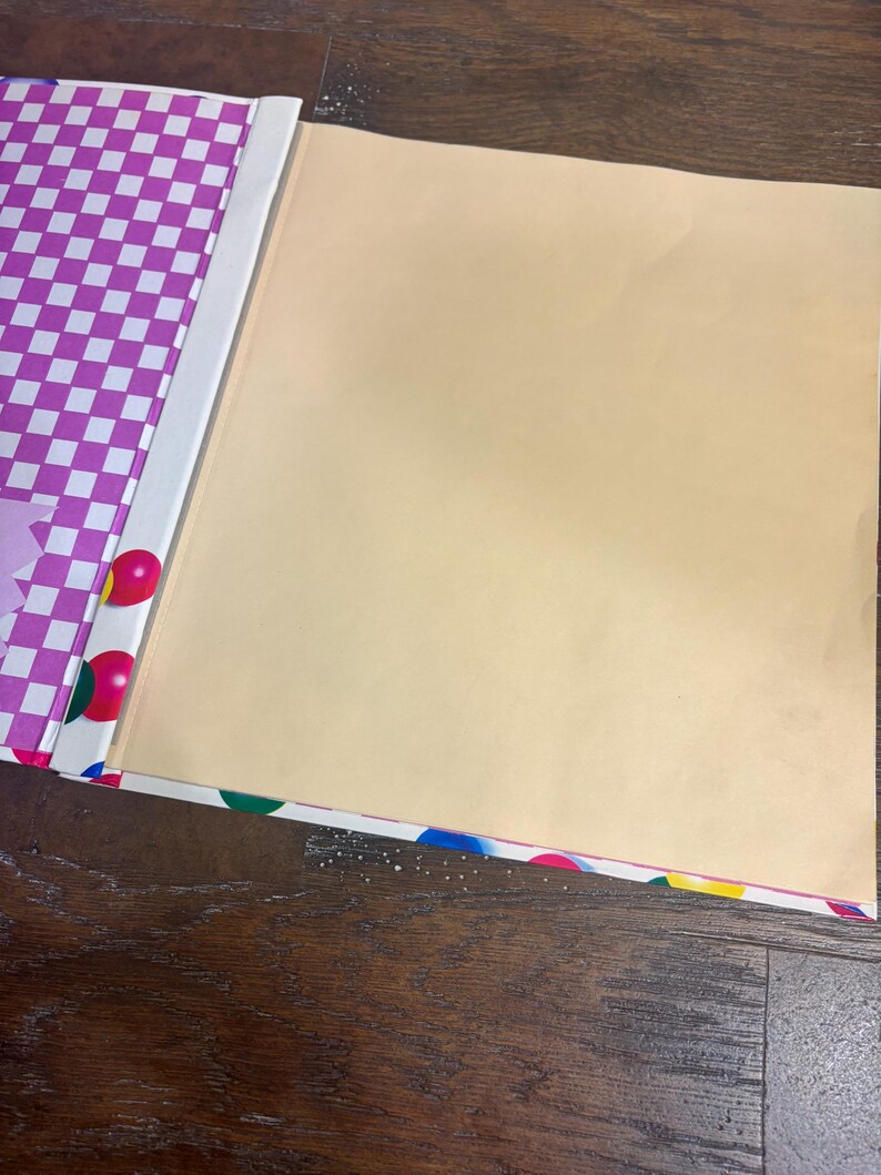 Puede incluir: Un &aacute;lbum de recortes abierto con un interior de color beige claro y una cubierta decorativa. La cubierta presenta un patr&oacute;n de cuadros morados y blancos, con lunares coloridos y un lomo blanco. El &aacute;lbum de recortes est&aacute; sobre una superficie de madera.