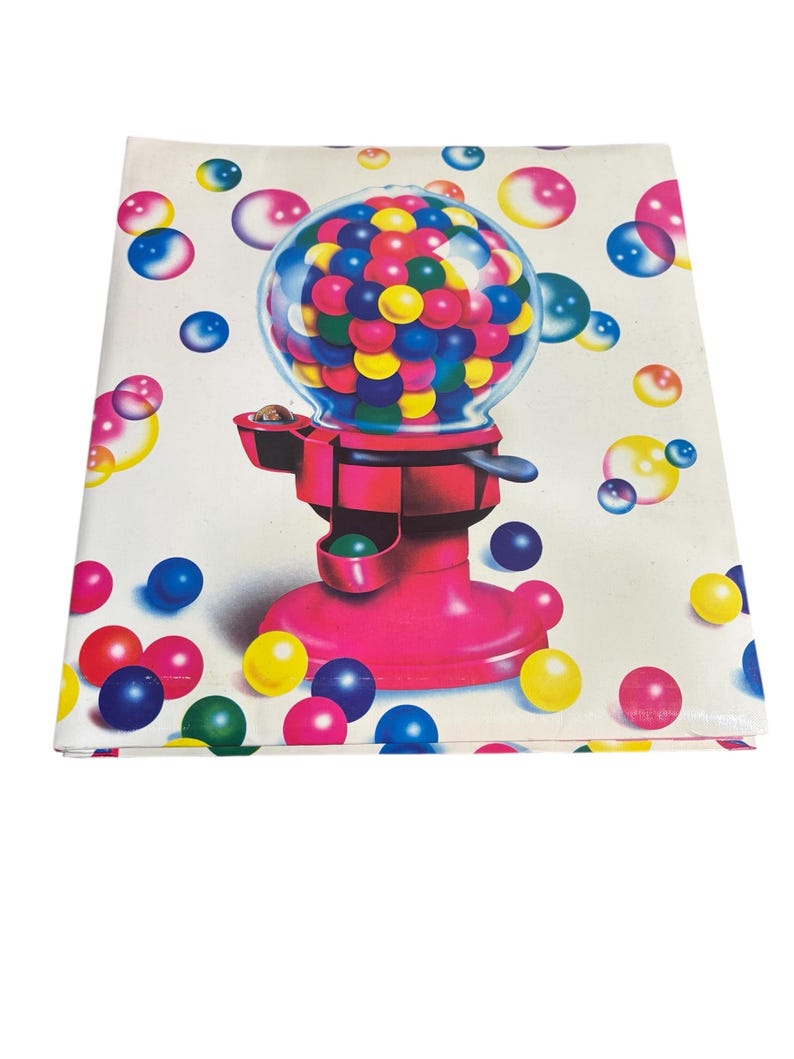 Puede incluir: Una tela blanca doblada con una ilustraci&oacute;n de una m&aacute;quina de chicles. La m&aacute;quina rosa tiene un globo transparente lleno de chicles de colores. Chicles de varios tama&ntilde;os en tonos azules, amarillos y rosas est&aacute;n dispersos.