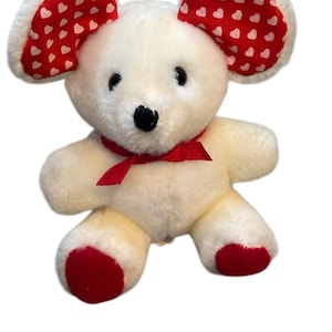 May include: A plush white mouse toy with large ears patterned with red hearts. The mouse has a red bow around its neck and red accents on its feet. The toy is designed to be soft and cuddly.
