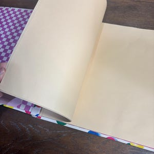 Puede incluir: Un cuaderno abierto con p&aacute;ginas color crema. La cubierta presenta un patr&oacute;n de cuadros morados y blancos, y el lomo est&aacute; decorado con lunares de colores. Las p&aacute;ginas se est&aacute;n pasando, revelando el interior en blanco.
