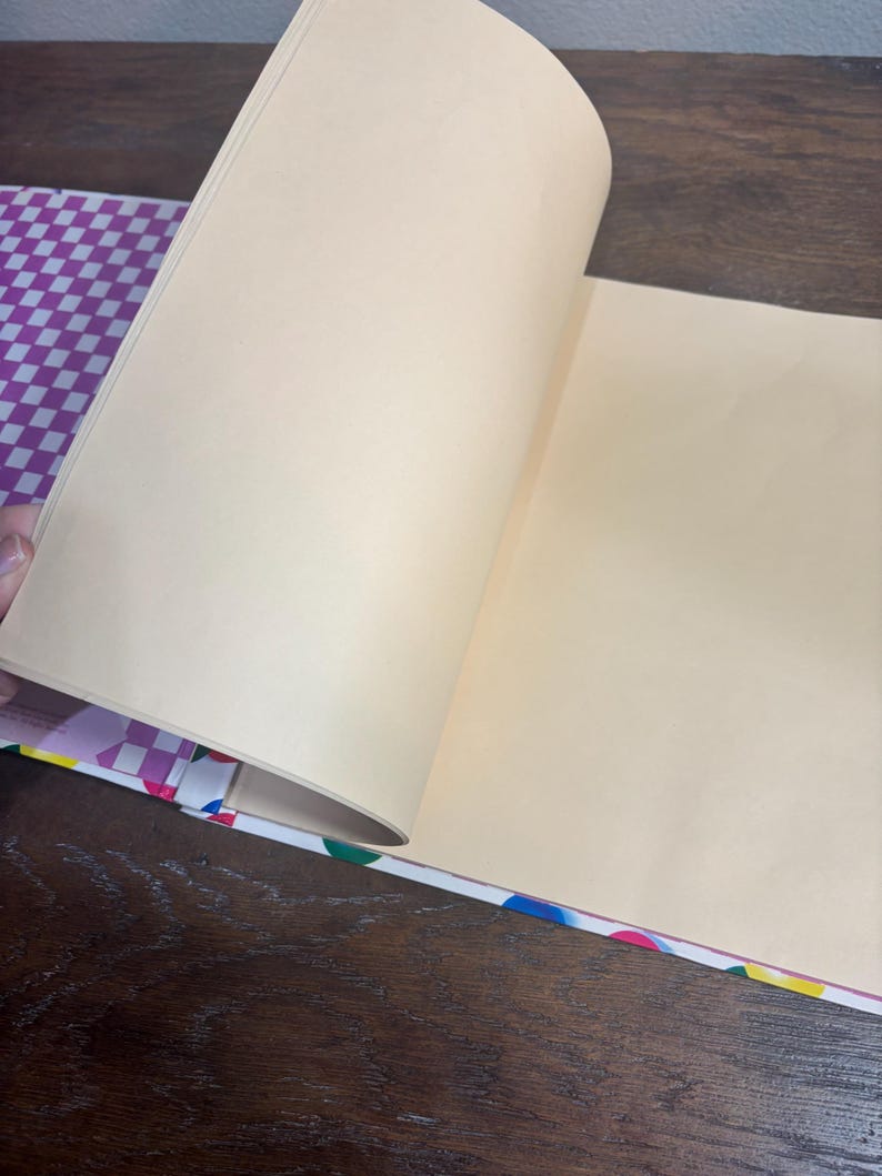 Puede incluir: Un cuaderno abierto con p&aacute;ginas color crema. La cubierta presenta un patr&oacute;n de cuadros morados y blancos, y el lomo est&aacute; decorado con lunares de colores. Las p&aacute;ginas se est&aacute;n pasando, revelando el interior en blanco.
