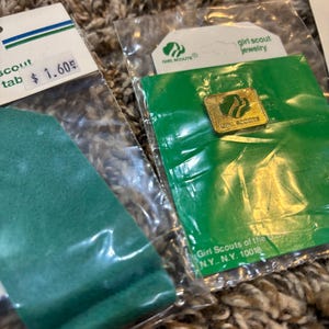 May include: Close-up of Girl Scout jewelry and a green scout tab in plastic packaging. The jewelry is a gold-colored pin with the Girl Scout logo. The scout tab has a price tag of $1.60.
