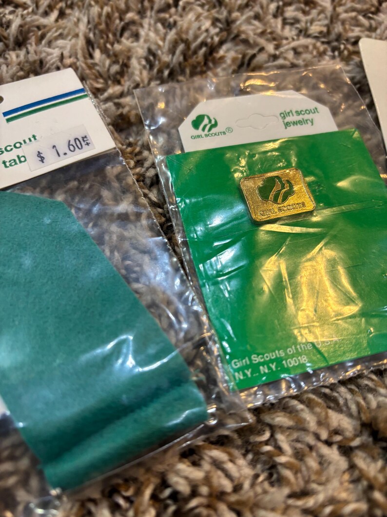 May include: Close-up of Girl Scout jewelry and a green scout tab in plastic packaging. The jewelry is a gold-colored pin with the Girl Scout logo. The scout tab has a price tag of $1.60.