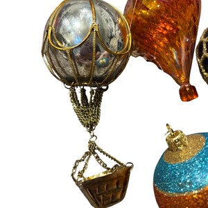 Może przedstawiać: Ozdobne szklane ozdoby choinkowe. Jedna ozdoba to srebrny balon na gorące powietrze ze złotą metalową ramą i koszem. Druga ma kształt łzy z pomarańczowymi i złotymi odcieniami. Trzecia jest niebiesko-złota.