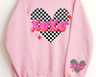 May include: Pink sweatshirt featuring a checkered heart design with the text "xoxo" in pink and red lettering. The heart has a white and black checkered pattern. The sweatshirt has a checkered heart design on the sleeve.