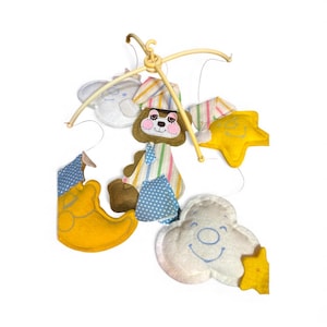 May include: A baby mobile featuring felt shapes of a bear, elephant, moon, star, and cloud. The bear has a striped hat and a blue bow tie. The moon and star are yellow. The cloud has a smiling face. The mobile is suspended from a plastic frame.