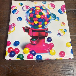 Puede incluir: Un cuaderno blanco con una ilustraci&oacute;n vibrante de una m&aacute;quina de chicles. La m&aacute;quina es de color rosa brillante con un globo transparente lleno de chicles de colores. Hay chicles flotantes de varios colores esparcidos.