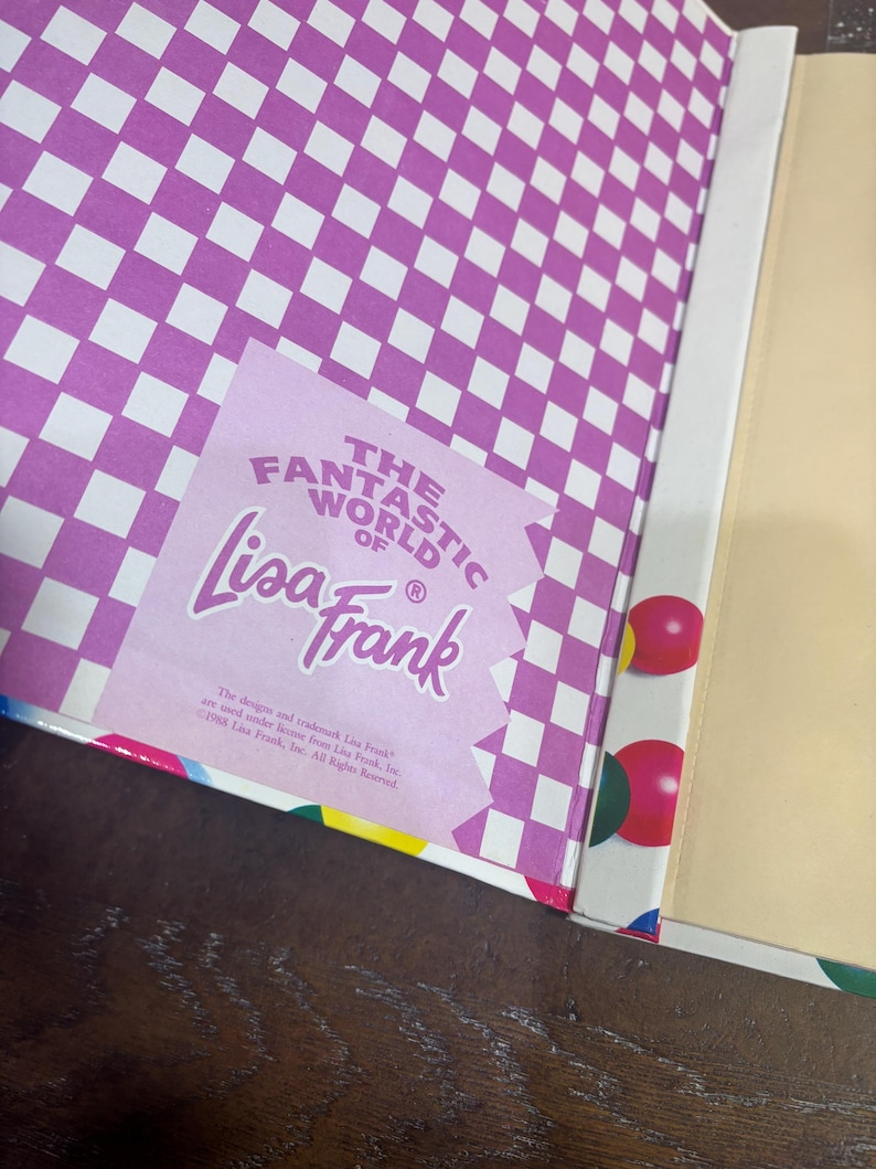 Puede incluir: Libro abierto con un patr&oacute;n de cuadros morados y blancos en la cubierta interior. Es visible una pegatina con el texto "The Fantastic World of Lisa Frank". El borde del libro tiene formas geom&eacute;tricas coloridas.