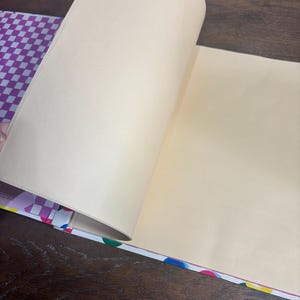 Puede incluir: Un cuaderno abierto con p&aacute;ginas de color crema. El lomo del cuaderno presenta un patr&oacute;n de lunares de colores, y la cubierta tiene un dise&ntilde;o a cuadros morado y blanco. Las p&aacute;ginas se est&aacute;n pasando, revelando el interior en blanco.