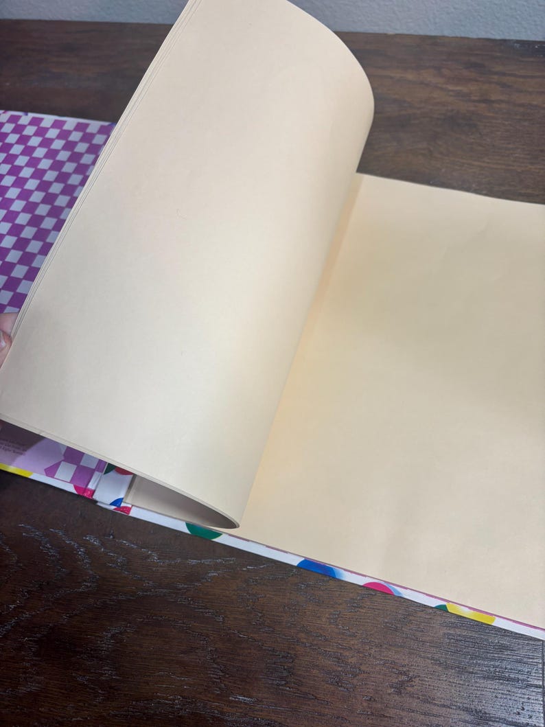 Puede incluir: Un cuaderno abierto con p&aacute;ginas de color crema. El lomo del cuaderno presenta un patr&oacute;n de lunares de colores, y la cubierta tiene un dise&ntilde;o a cuadros morado y blanco. Las p&aacute;ginas se est&aacute;n pasando, revelando el interior en blanco.