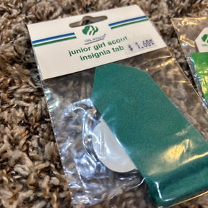 May include: A green Junior Girl Scout insignia tab in a clear plastic package. The package has a white and green label with the Girl Scouts logo and the text "junior girl scout insignia tab".