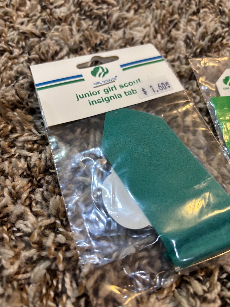 May include: A green Junior Girl Scout insignia tab in a clear plastic package. The package has a white and green label with the Girl Scouts logo and the text "junior girl scout insignia tab".