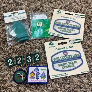 May include: A collection of Girl Scout patches and insignia, including a Junior Girl Scout insignia tab, Council ID sets, and various embroidered patches with numbers and community service themes. The items are displayed on a textured brown surface.