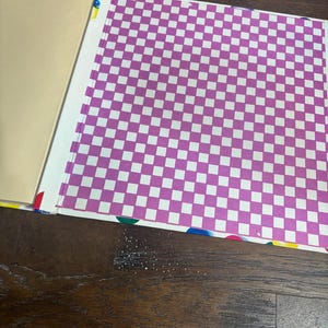 Puede incluir: Cuaderno abierto con una cubierta beige y una p&aacute;gina con un patr&oacute;n de cuadros morados y blancos. La encuadernaci&oacute;n es visible con acentos coloridos. El cuaderno est&aacute; sobre una superficie de madera oscura.