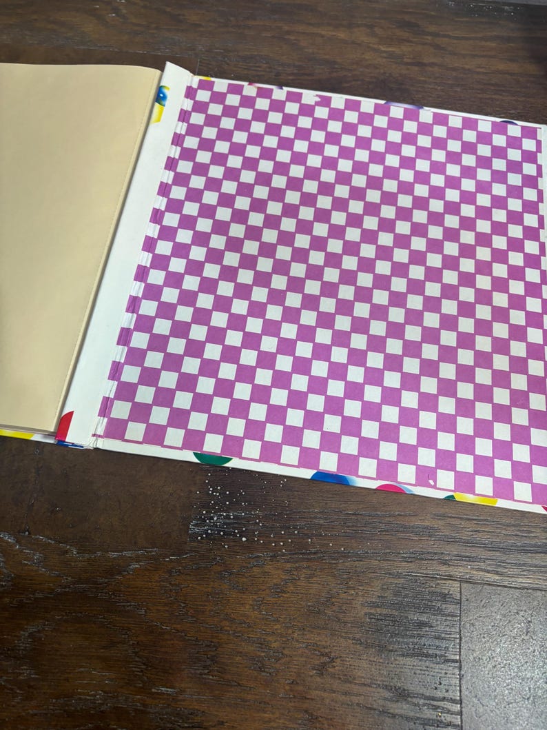 Puede incluir: Cuaderno abierto con una cubierta beige y una p&aacute;gina con un patr&oacute;n de cuadros morados y blancos. La encuadernaci&oacute;n es visible con acentos coloridos. El cuaderno est&aacute; sobre una superficie de madera oscura.