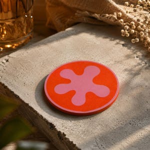 Puede incluir: Un posavasos redondo con un fondo naranja brillante y un diseño floral abstracto rosa en el centro. El posavasos está hecho de un material similar al fieltro y se encuentra sobre una superficie de hormigón. Diámetro: aprox. 10 cm.