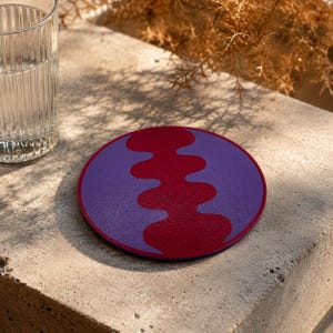 Pode incluir: Uma base de copo redonda com fundo roxo e um design ondulado bordô. A base de copo está sobre uma superfície de concreto ao lado de um copo de água. A base de copo é um item decorativo, com cerca de 10 cm de diâmetro.