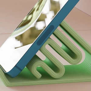 Può includere: Un supporto per telefono verde chiaro con un design curvo unico. Sostiene uno smartphone blu, angolato per la visualizzazione. Il design del supporto presenta una serie di supporti a coste ad anello, che offrono un'estetica moderna. Lo sfondo è di colore neutro.