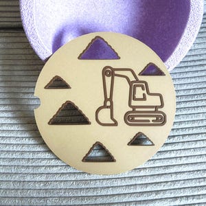 Può includere: Un sottobicchiere beige per auto con una ruspa marrone e disegni a triangolo marrone. Il sottobicchiere rotondo ha una piccola tacca. Lo sfondo è un contenitore viola chiaro e una superficie grigia strutturata.