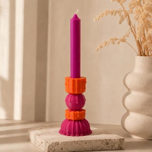 Op de afbeelding: Een levendige, gestapelde kandelaar met een magenta kaars. De houder bestaat uit oranje en magenta geometrische vormen. De kandelaar staat op een stenen oppervlak, met een vaas met gedroogde bloemen op de achtergrond.