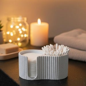 Può includere: Un organizer da bagno grigio chiaro e scanalato con due scomparti. Un lato contiene dischetti di cotone, l'altro cotton fioc. Una candela accesa e un barattolo di lucine sullo sfondo suggeriscono un'atmosfera da spa.
