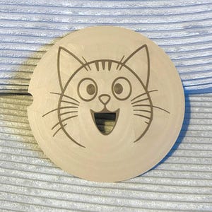 Può includere: Una placca rotonda in legno con una finitura marrone chiaro presenta un design di gatto dei cartoni animati inciso a laser con occhi grandi, un ampio sorriso e baffi.