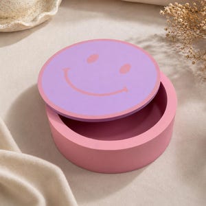 Könnte beinhalten: Eine runde, rosafarbene Aufbewahrungsbox mit abnehmbarem Deckel. Der Deckel ist hellviolett und zeigt ein einfaches, rosafarbenes Smiley-Gesicht, das aus zwei ovalen Augen und einem geschwungenen Mund besteht, der mittig auf dem Deckel liegt.