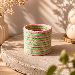 Könnte beinhalten: Ein zylinderförmiger Behälter mit abwechselnd horizontalen rosa und grünen Streifen. Der Behälter besteht aus einem gewebten Material und steht auf einer Betonoberfläche. Der Hintergrund umfasst eine Vase mit getrockneten Blumen und ein Holztablett mit zwei Eiern.