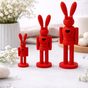 Pode incluir: Três estatuetas de coelhos vermelhos de tamanhos variados. Cada um tem orelhas compridas, um laço e um recorte em forma de coração. Os coelhos estão em bases redondas e são exibidos em uma superfície clara.