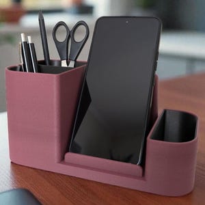Könnte beinhalten: Ein bordeauxfarbener Schreibtisch-Organizer mit Fächern für Stifte, Bleistifte, Scheren und ein Smartphone. Der Organizer besteht aus einem festen Material und steht auf einer Holzoberfläche. Das Telefon ist schwarz.