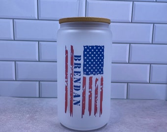 May include: White tumbler with a red, white, and blue American flag design. The flag has a distressed look and the name "BRENDAN" is written vertically down the left side of the flag. The tumbler has a bamboo lid and a clear plastic straw.