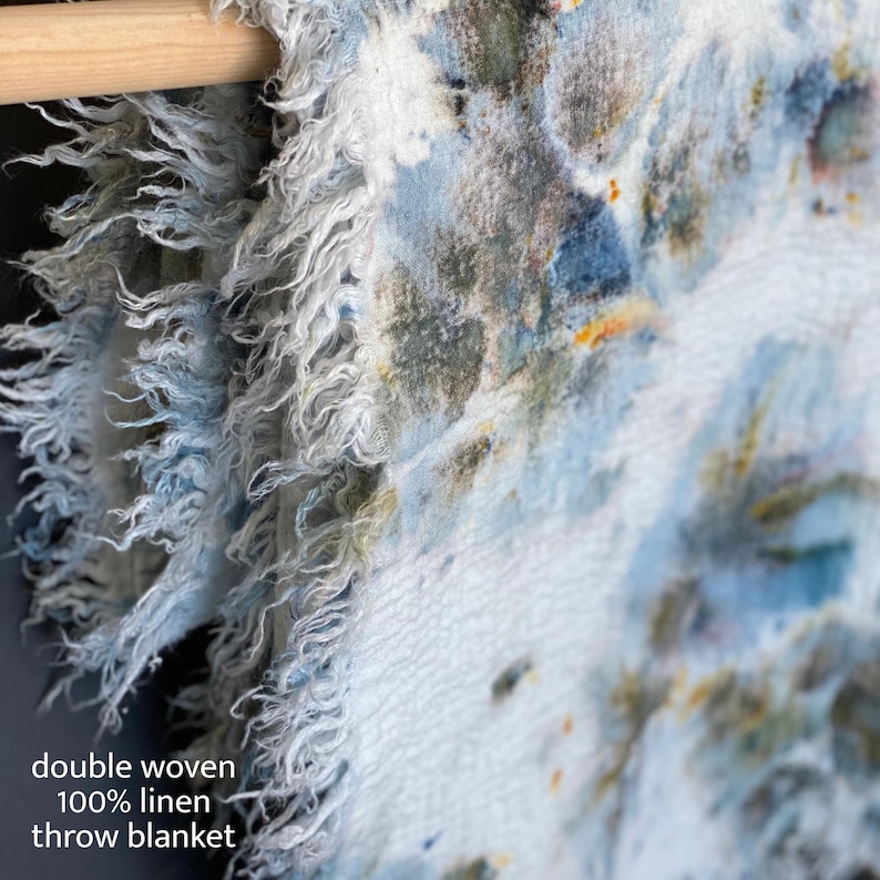 May include: A double woven 100% linen throw blanket with a watercolor design in shades of blue, white, and touches of yellow and green. The blanket has a fringed edge and is hanging on a wooden rod.