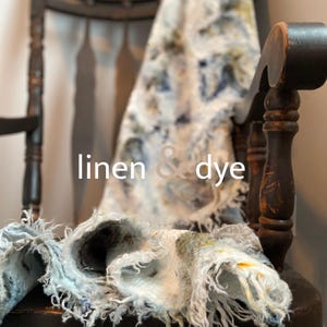 May include: A close-up of a textile with the words "linen & dye" in white. The textile is draped over a dark wooden chair. The textile has a blue, white, and yellow pattern. The textile has a frayed edge.