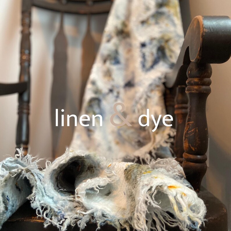 May include: A close-up of a textile with the words "linen & dye" in white. The textile is draped over a dark wooden chair. The textile has a blue, white, and yellow pattern. The textile has a frayed edge.