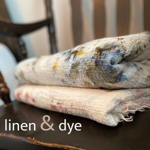 May include: Two folded linen textiles with a textured appearance. The top textile features a pattern of blue, yellow, and green hues, while the bottom one has pink and red accents. The text "linen & dye" is at the bottom.